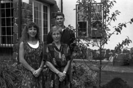 Julie Anderson, Suzie Watts, and Jeffrey Watts stand next to the tree dedicated to Brady Watts near Lewis House (1973), St. Cloud State University