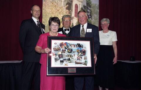 Color image of five people in formal attire stand in front of a red curtain backdrop. The woman and man in the center holds a large framed collage of photographs. A decorative cupola with greenery is partially visible behind them.