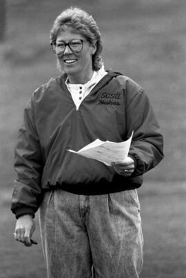Black and white image of a woman standing outdoors in a grassy area, wearing glasses, a light-colored shirt, jeans, and a jacket with “SCSU Huskies” on it, holding papers in one hand.