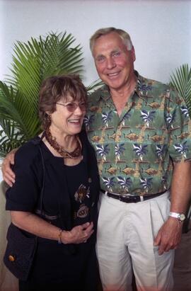 Color image of a man and a woman standing together in front of green palm leaves. The man wears a green tropical-print shirt and beige pants, with his arm around the woman’s shoulders. The woman wears glasses and a black outfit with colorful patterns, holding a purse.