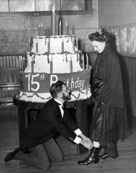 Harry Schuelke ties the shoe of Lois Nissen in front of large birthday cake, St. Cloud State University